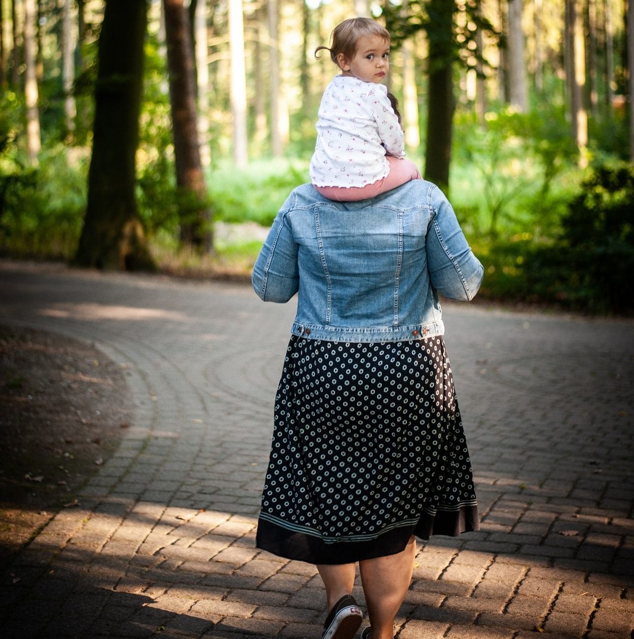 A woman in a denim jacket and polka dot skirt carrying a toddler on her shoulders in a sunlit forest.
