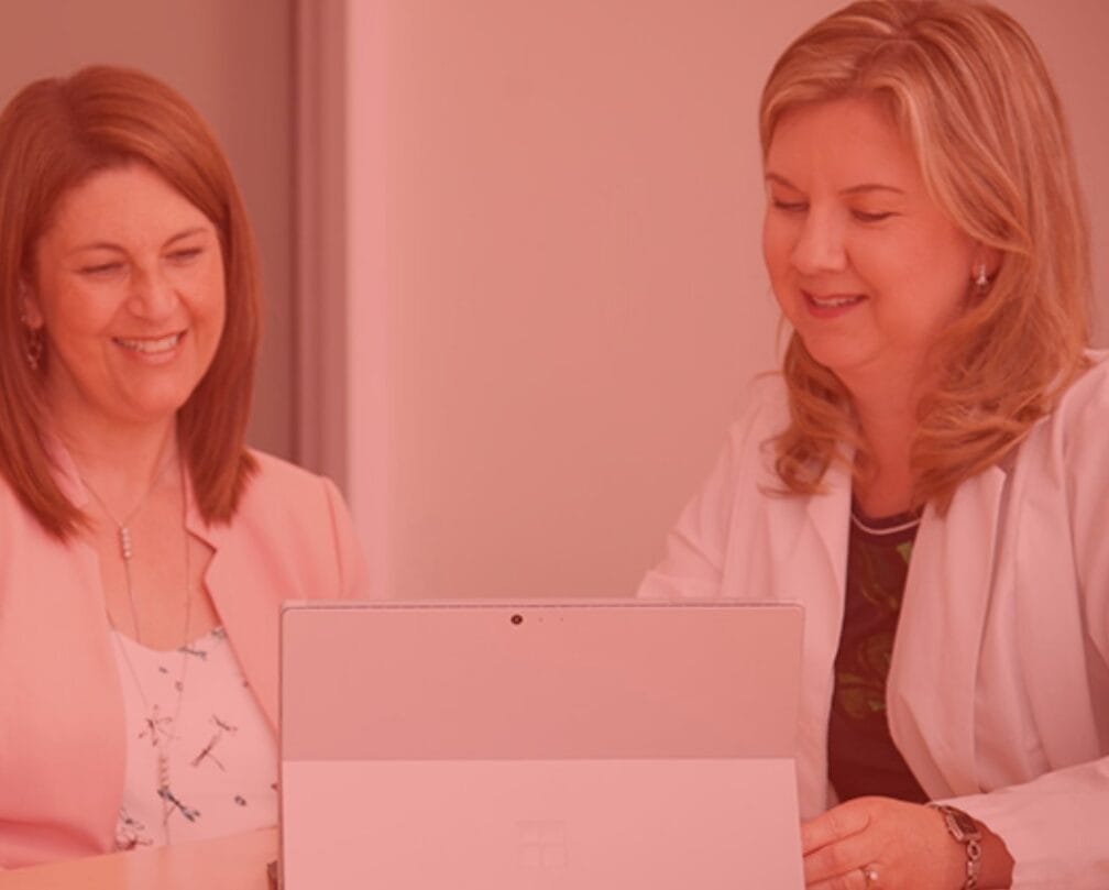 Two women, one viewing a laptop screen with a smile, in a room with a pink hue.