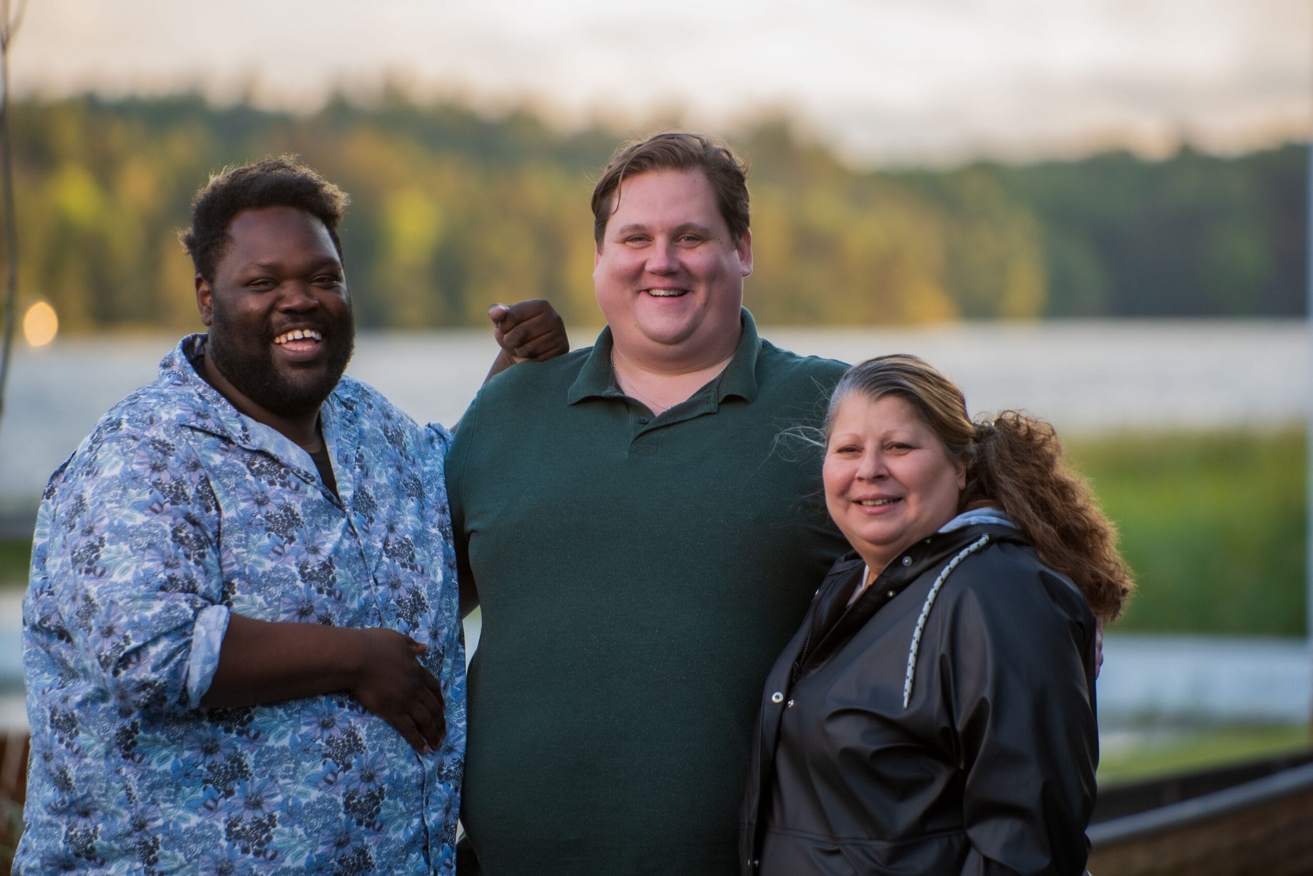 Three friends smiling together by a lake at sunset, with trees and water in the background. .
