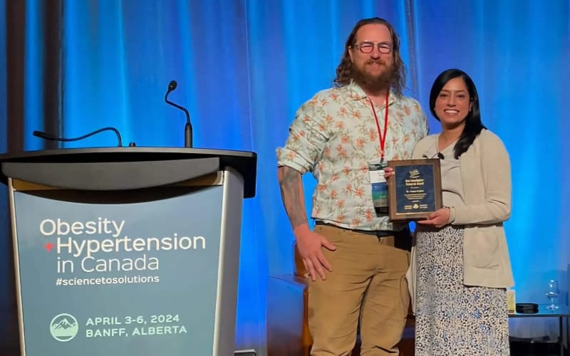 Two individuals standing beside a podium at a conference, holding an award plaque, with a banner reading "obesity + hypertension in canada" in the background.