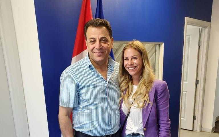 Two people standing side by side, smiling in front of a white wall with two flags behind them, one red and white, the other blue. One wears a striped shirt and dark pants, the other a purple blazer and jeans.